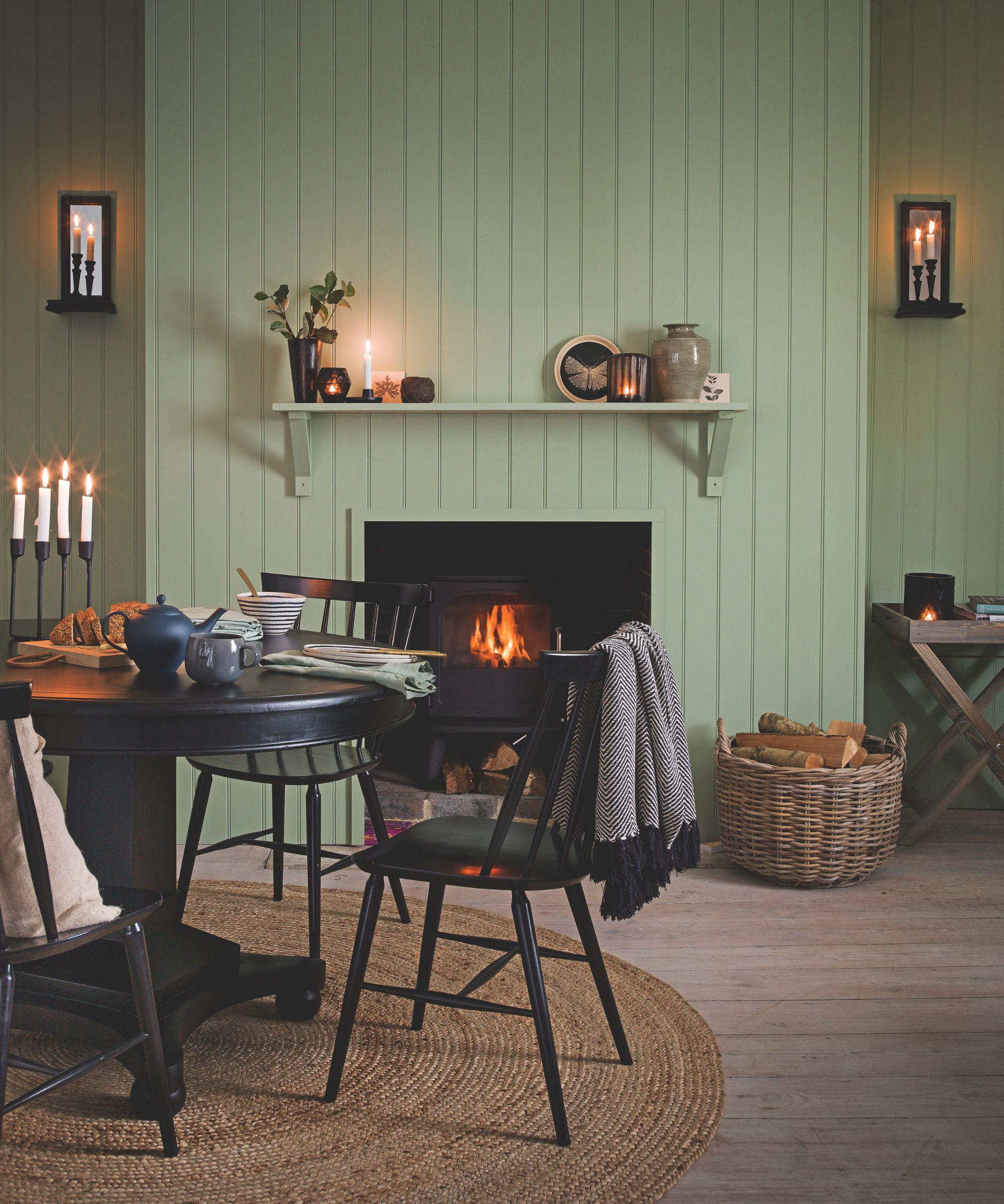 A green panelled living room with a lit fire in the fireplace, rattan circular rug, circular black dining table with matching chairs, a wicker basket of firewood and lit taper candles.
