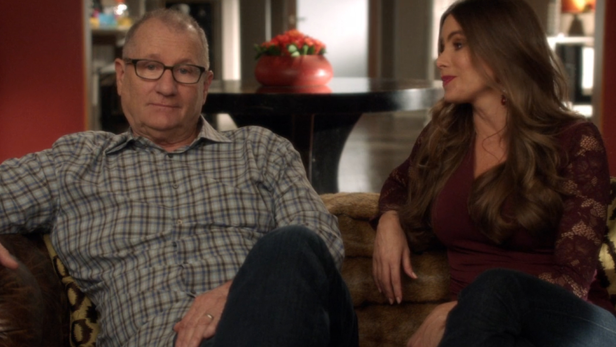 Jay and Gloria sit on the couch together talking to the camera in Modern Family.