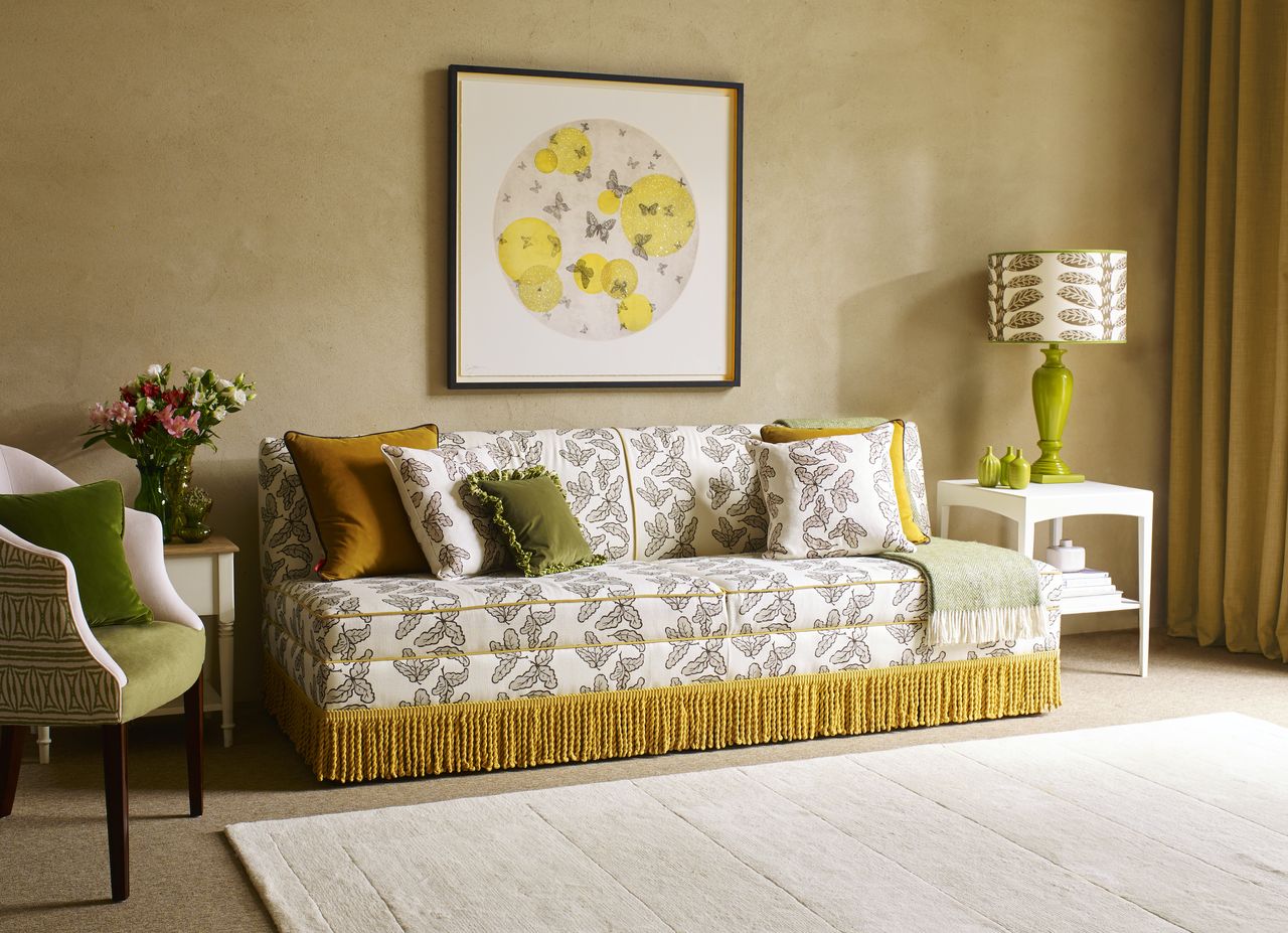 beige living room with patterned sofa with fringing, greensand blush armchair, oatmeal carpet, turmeric drapes, white side table, green table lamp base and patterned shade, bee artwork, cream rug