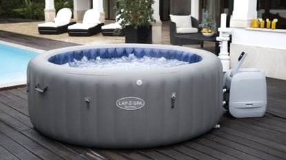 A grey inflatable Lay-Z-Spa hot tub on decking by a pool with sun loungers in the background