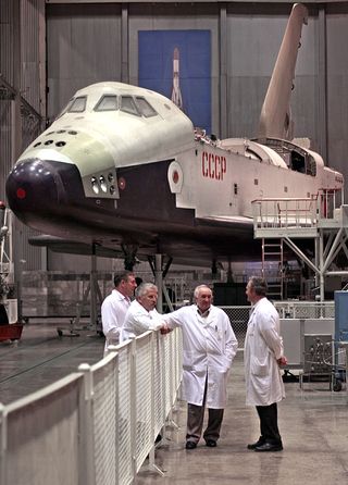 four men in white lab coats stand at a fence in a large hangar with a space shuttle behind them.