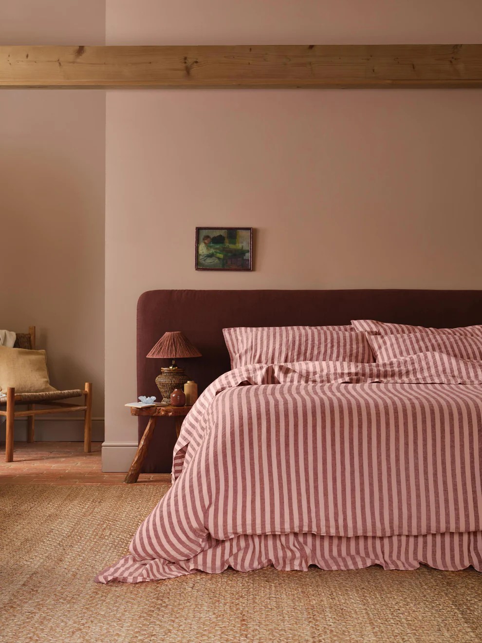 Image of a dusty pink bedroom with a jute rug and a large bed in the center. The bed has an aubergine purple headboard with bedding that is pink and berry-colored stripes.