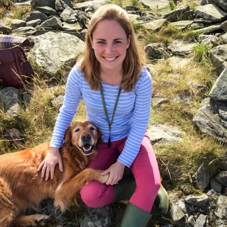 Dr Aimee Warner is a white women, with shoulder length dark blonde hair. She is pictured outdoors, smiling, sat on mossy rocks with a very happy dog which is resting its paw on her leg.