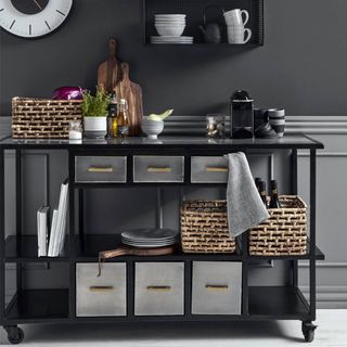 Portable black kitchen island with 6 grey draws and open shelves, set against a wall with dark grey paint at the top and light grey at the bottom