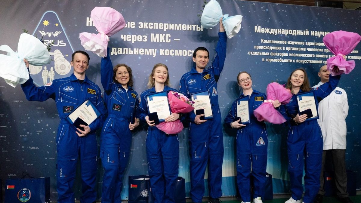 six people in blue flight suits wave in front of a blue-postered wall with text in russian on it