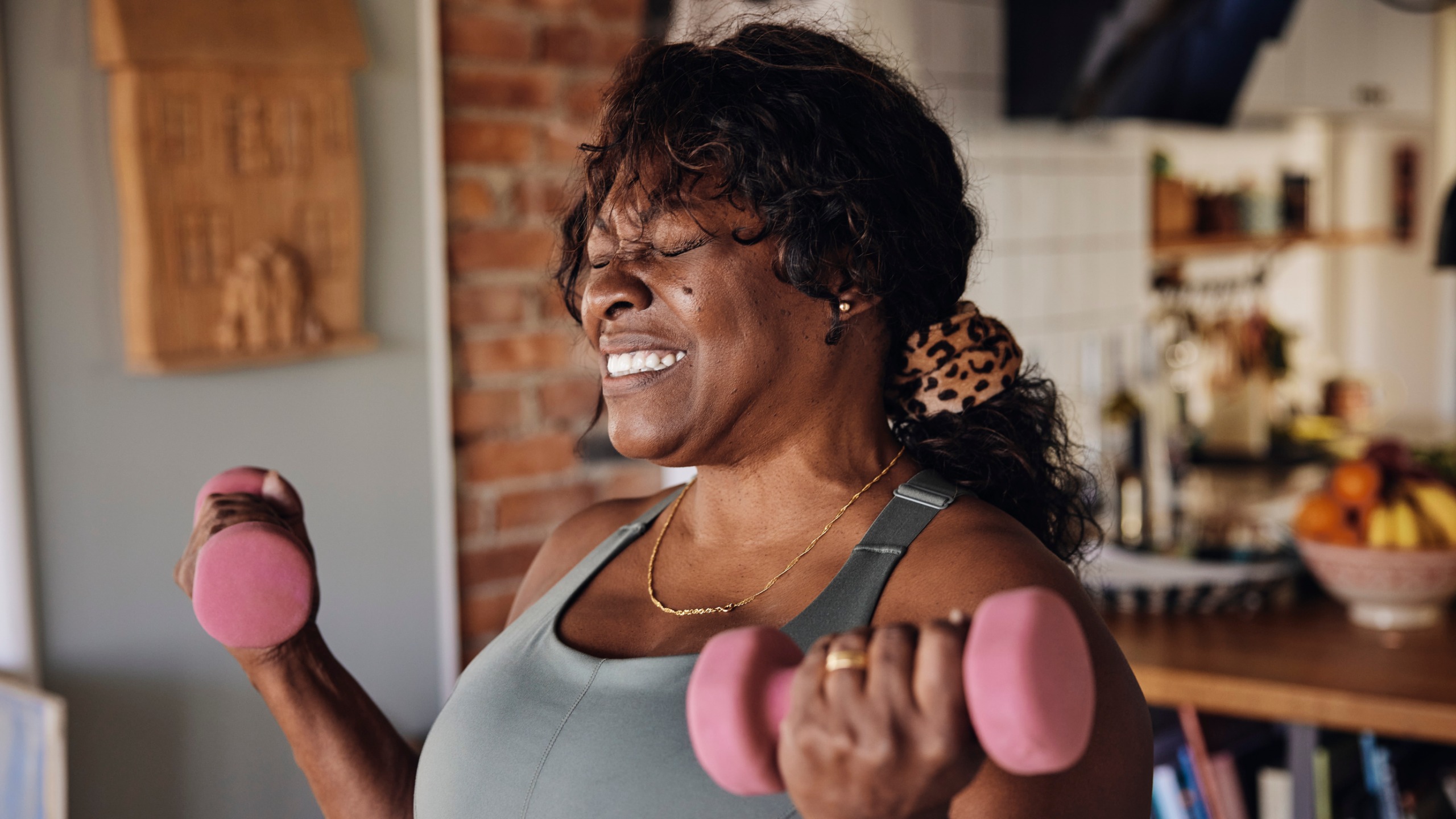 A woman grimaces as she lifts lightweight dumbbells up to her shoulders standing in her kitchen living room.