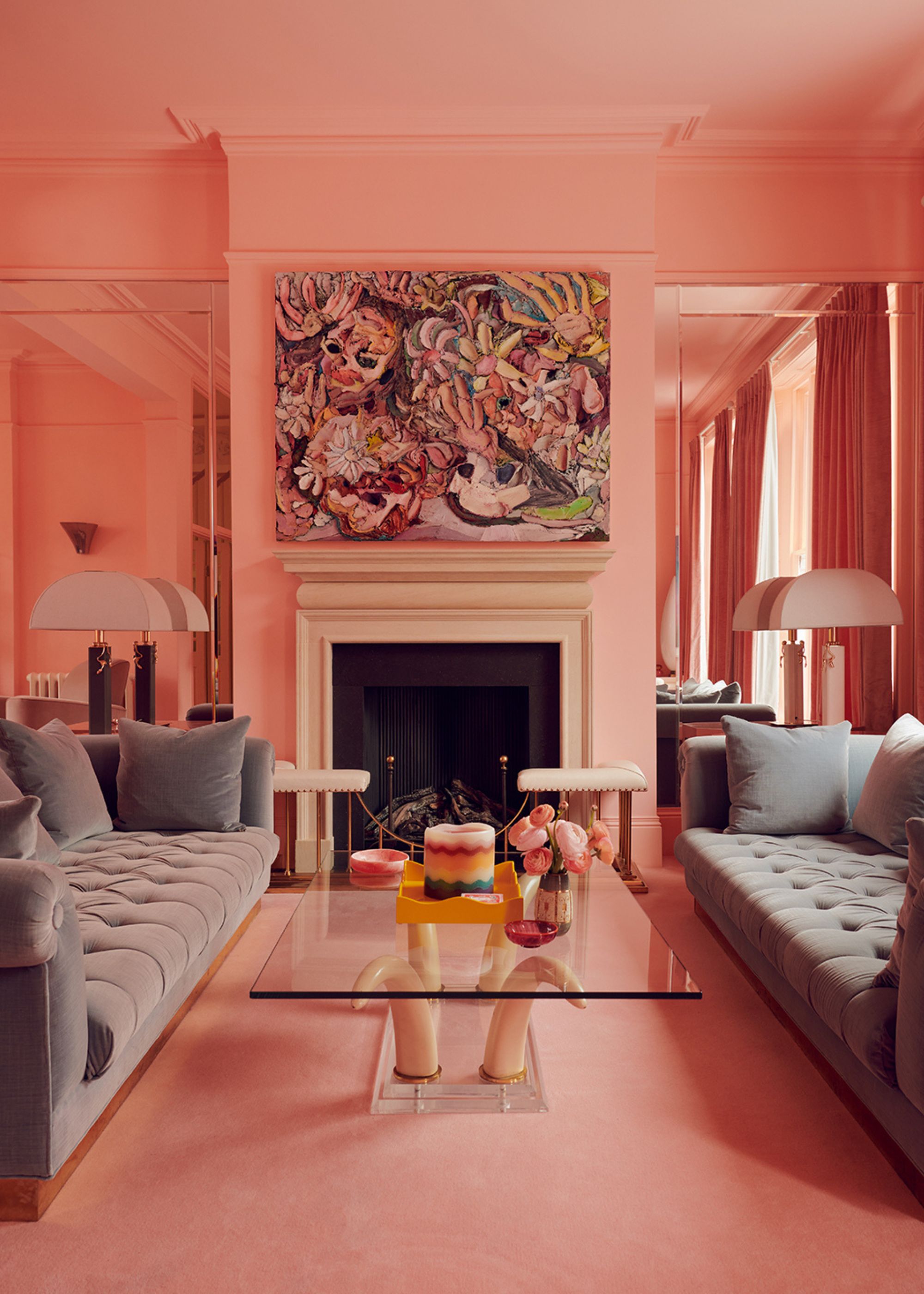 A modern pink living room with two gray sofas facing each other with a square glass coffee table in the center in front of a fireplace with artwork above it and large white table lamps either side