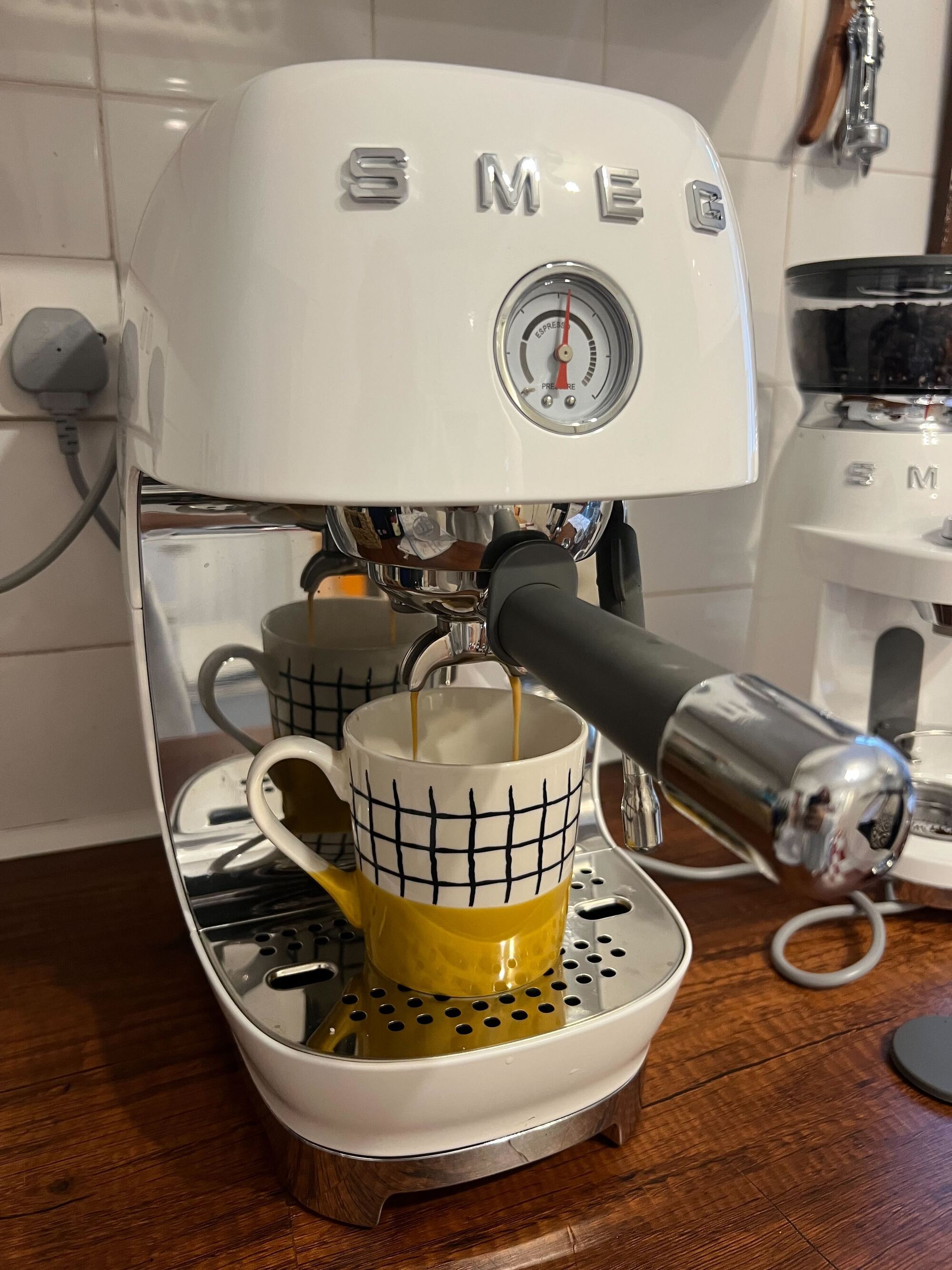 Image of a white Smeg cold brew machine brewing a shot of espresso on a wooden countertop with white tile backsplash