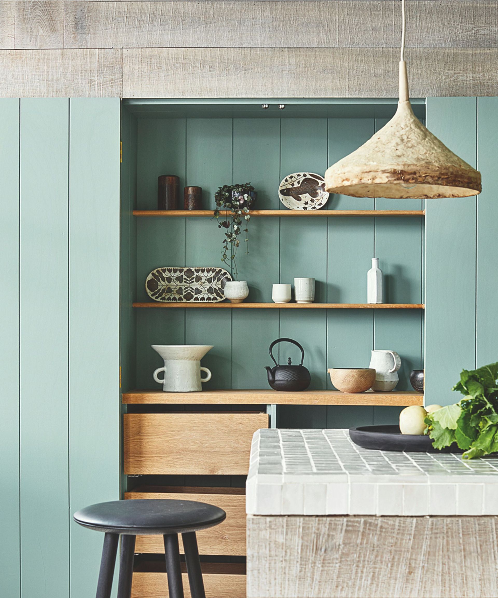 Duck egg blue wooden kitchen shelving with neatly arranged ceramic pots on, a wooden stool in front and a mosaic-topped kitchen island in the foreground