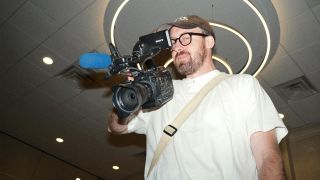 John Wilson, in a cap and white t-shirt, holds a camera on his shoulder in How To with John Wilson