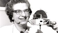 a woman wearing thick rim glasses holds a scale model of a satellite in this black and white photo