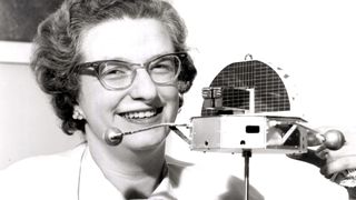 a woman wearing thick rim glasses holds a scale model of a satellite in this black and white photo