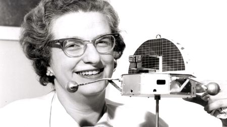 a woman wearing thick rim glasses holds a scale model of a satellite in this black and white photo