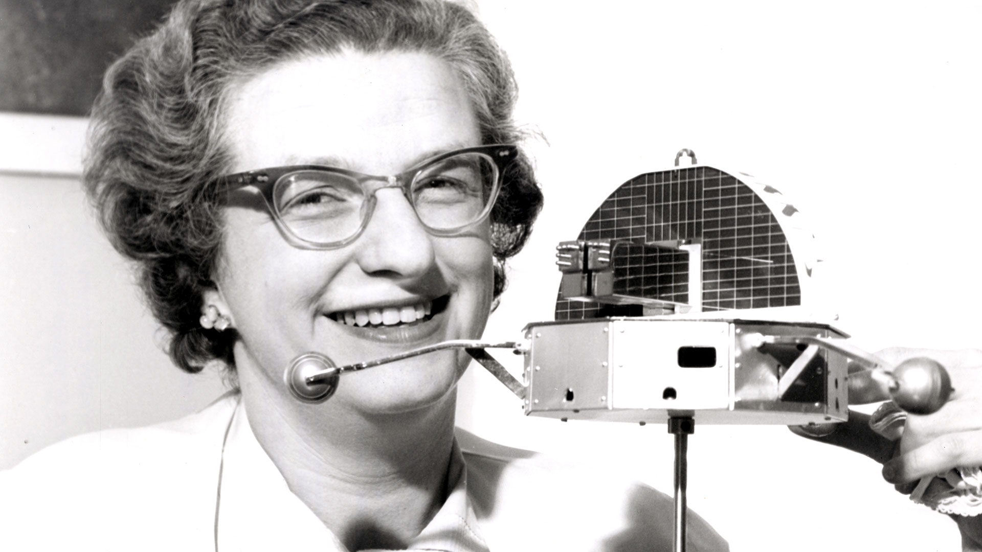 a woman wearing thick rim glasses holds a scale model of a satellite in this black and white photo