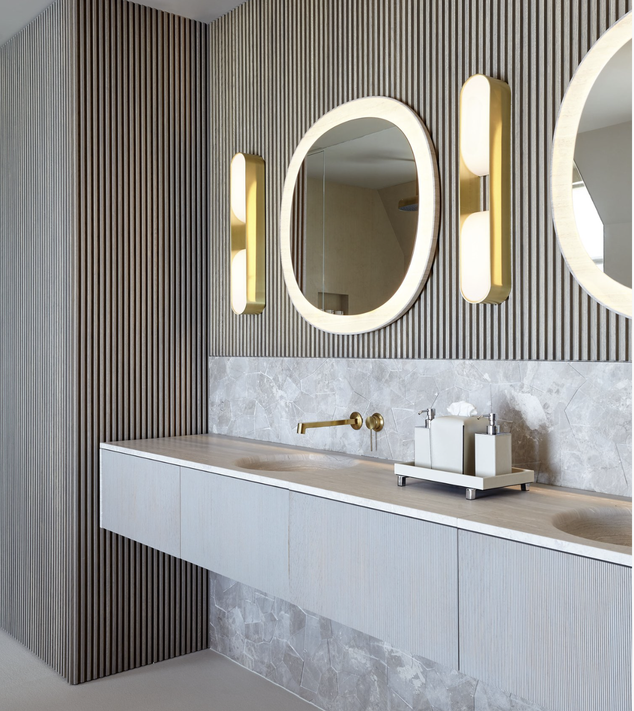 The interiors of a bathroom features modern furnishings including neon-lit mirrors in curvilinear shapes, accent walls in wood and stone, and chrome finishes.
