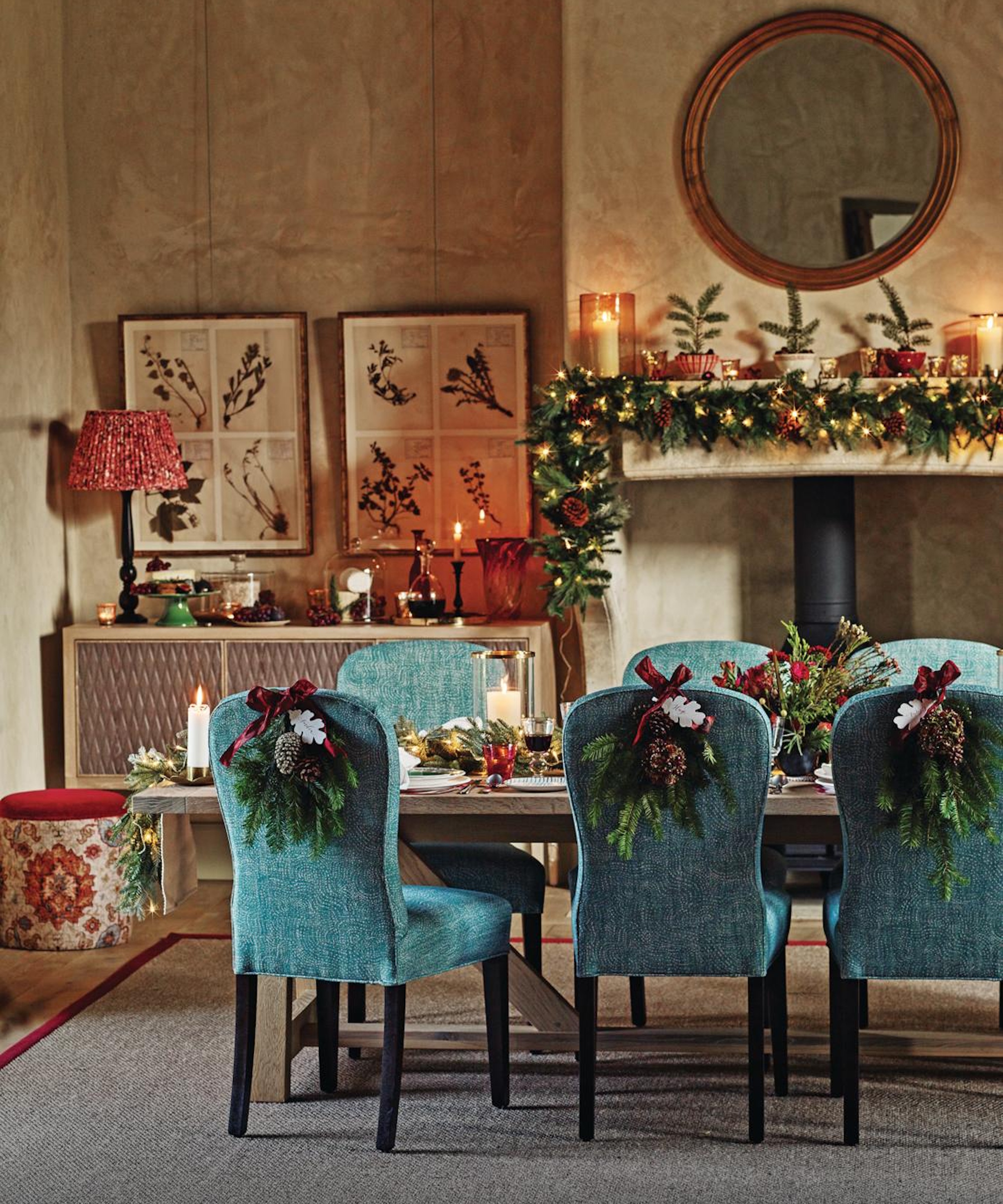 Christmas table with blue seats, each one with a wreath on the back. A garland sits on the mantel with a round mirror above.