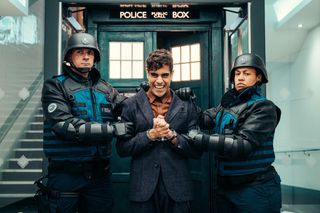 The Master (Sacha Dhawan) stands outside the Tardis. He is grinning evilly, and is in handcuffs, being restrained by a UNIT officer on either side
