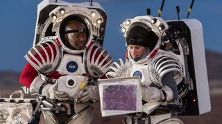 a man and woman in white spacesuits study maps in a desert landscape during a training exercise