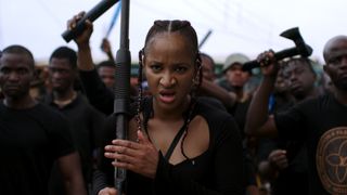 A still from the movie Gangs of Lagos in which one of the characters looks ahead at the camera while holding a gun and a group of people waving weapons stands behind her.