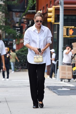 Gigi Hadid berjalan di NYC mengenakan kemeja berkancing putih dengan celana panjang hitam dan flat serta tas Loro Piana Extra Pocket.