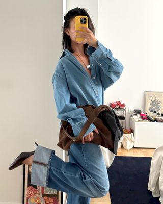 fashion influencer Sasha Mei poses for a mirror selfie wearing a denim button-down shirt, black cord necklace with a silver pendant, brown suede tote bag, cuffed jeans, and black pumps