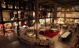 Opening dinner takes place in the industrial warehouse. Surrounding the cement floored atrium, are three floors of black metal balconies. Throughout the atrium, dinner tables are arranged. Some tables are round, some are long and rectangular.