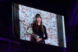 A photo of Taylor Swift delivering a virtual acceptance speech in a black long-sleeved t-shirt paired with pops of bright red on her lips and fingertips.