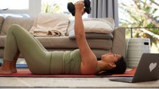 woman lying down holding two light dumbbells overhead living room setting in front of laptop