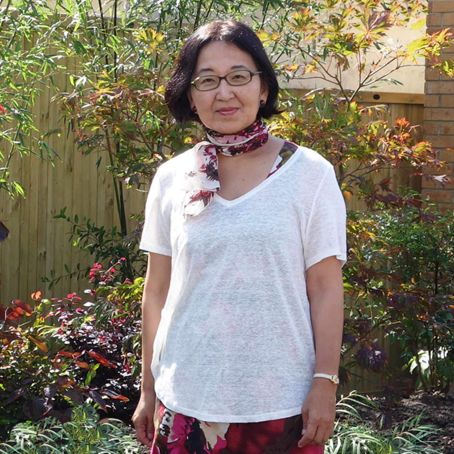 A woman with short black hair and a patterned neck scarf in a white v-neck and a patterned skirt in a garden