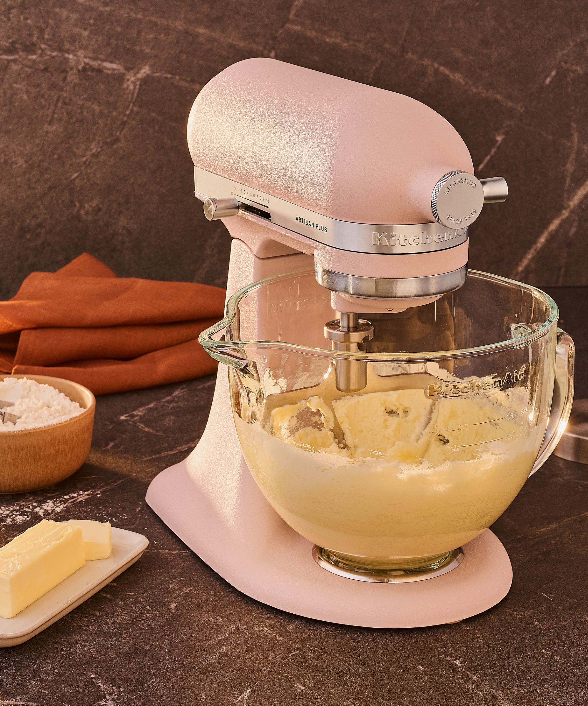 A pink KitchenAid Artisan Plus Stand Mixer with a clear bowl on a kitchen counter surrounded by baking ingredients.