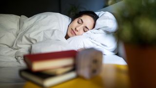 A woman sleeps peacefully in her bed under a white duvet, while a small stack of books can be seen on her bedside table