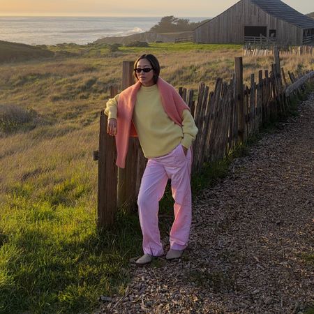 Aimee Song wears a pastel yellow sweater with a coral sweater draped over her shoulders and pink elastic waist pants in front of a sunset. 