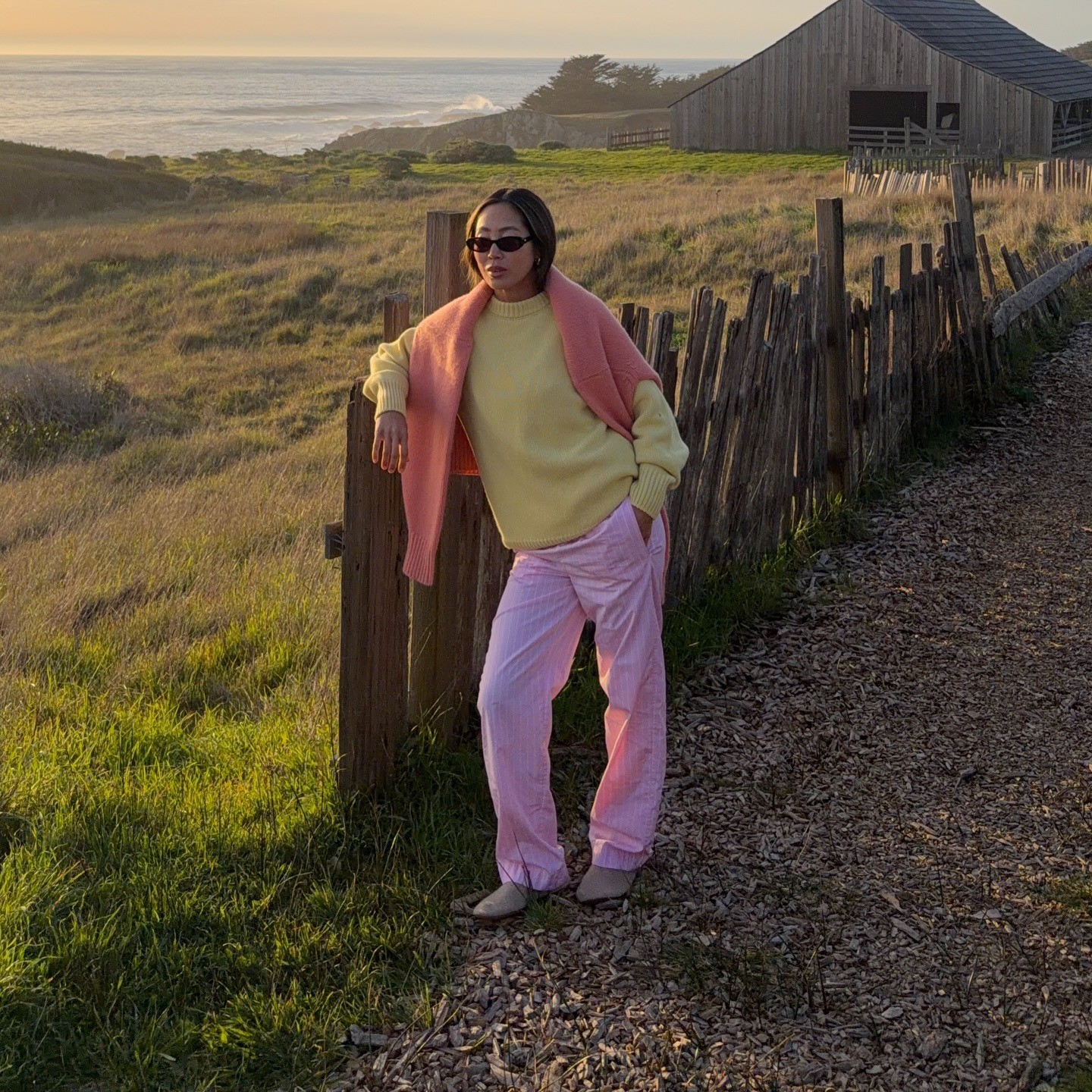 Aimee Song wears a pastel yellow sweater with a coral sweater draped over her shoulders and pink elastic waist pants in front of a sunset. 