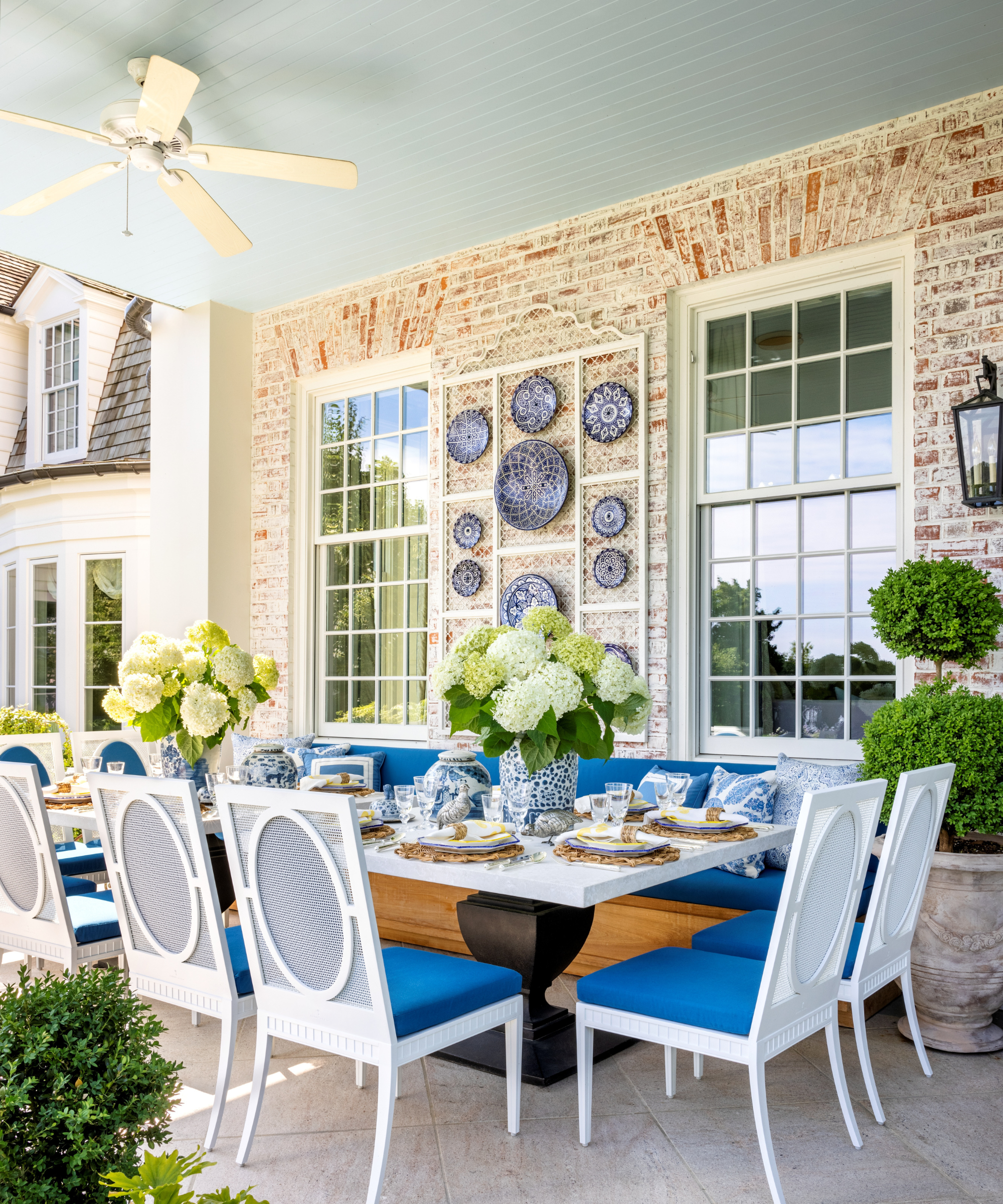 Outdoor pedestal dining space featuring layered plates, blue-cushioned chairs, green hydrangeas, and antique-style lanterns and plates mounted to its exterior brick wall.