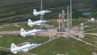 four jets at the left of a launch pad. there is a rocket on the launch pad with towers surrounding it
