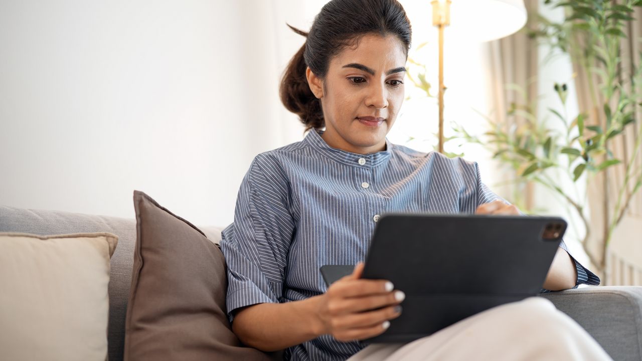 A woman sits on her sofa and looks at her tablet.