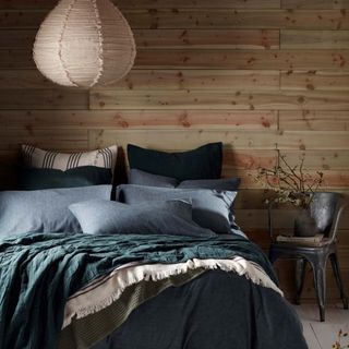 A bedroom with a timber clad wall and a bed covered in bedding in various shades of green