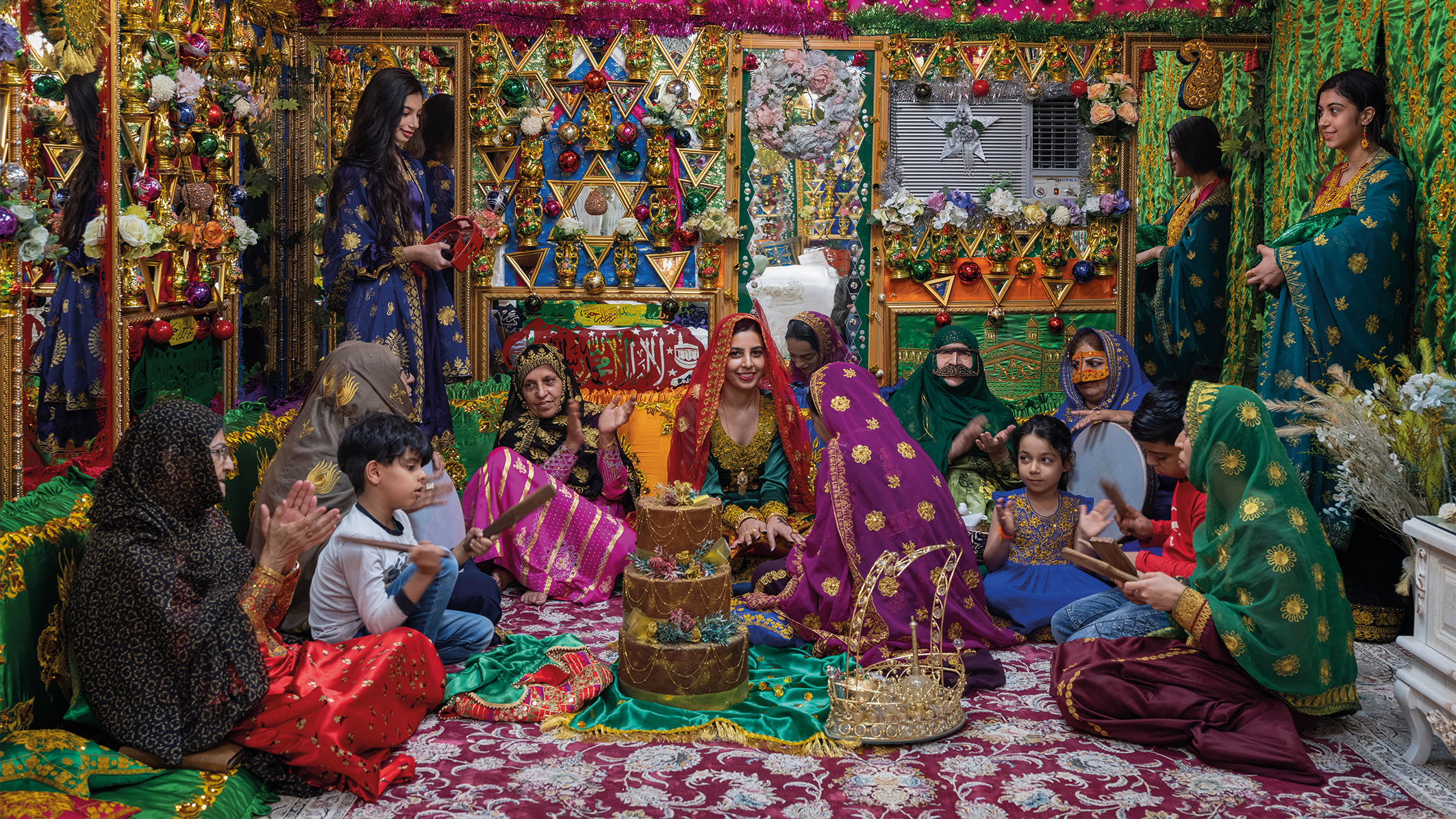 A vibrant celebration scene shows women and children in colorful traditional attire amidst ornate decorations, flowers, and a cake