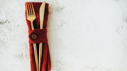 Gold coloured knife and fork laid on a burgandy napkin on a white marble background