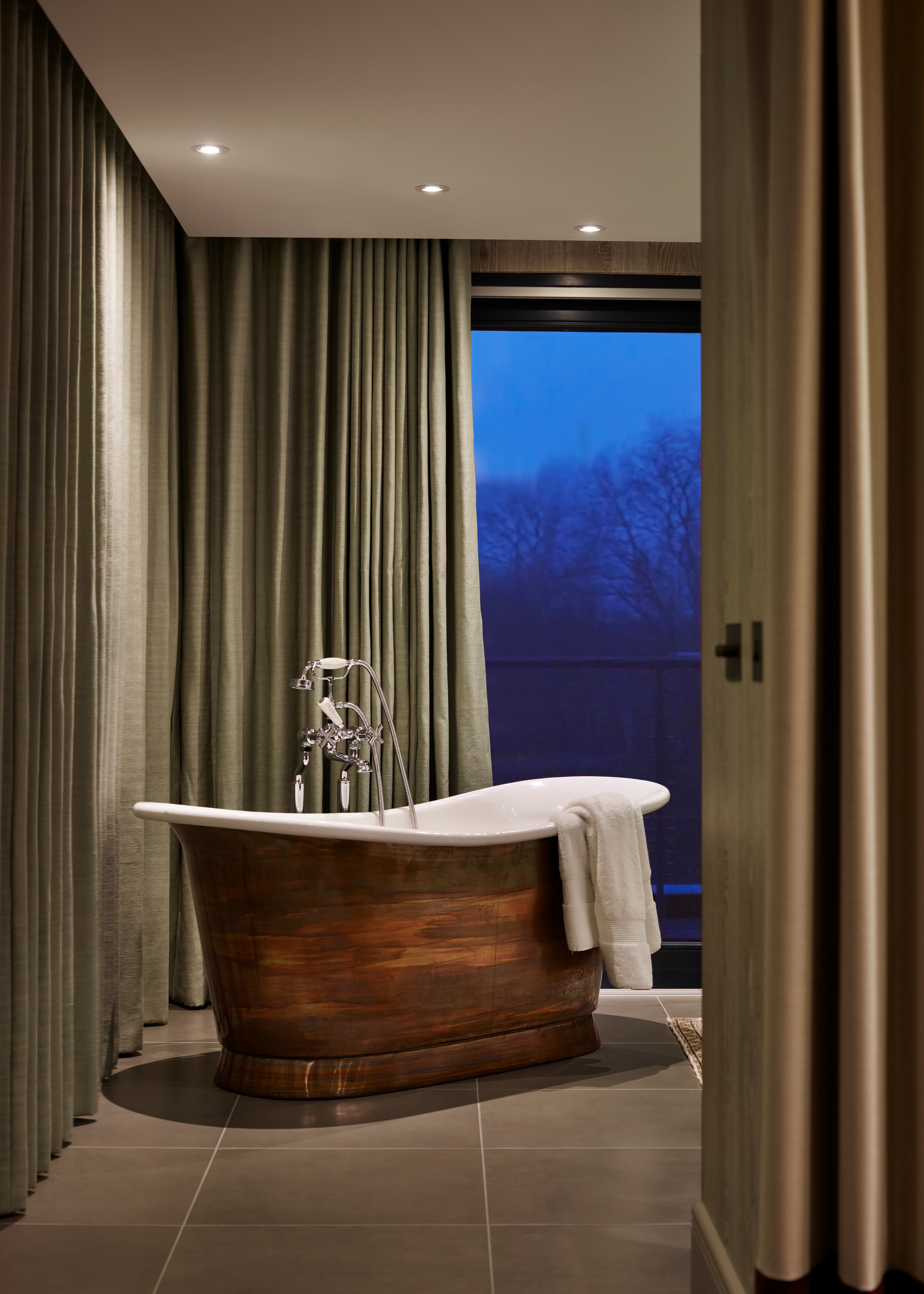 copper freestanding bath in the corner of a large low lit bathroom