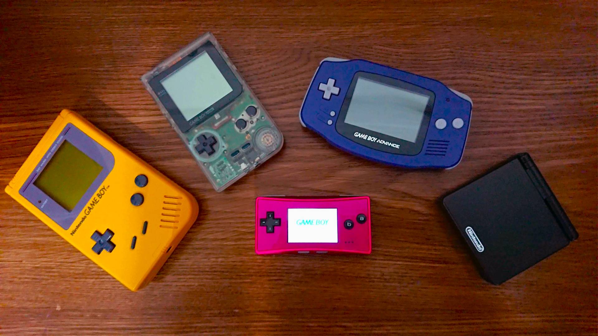 Game Boy models from left to right, starting with Yellow DMG, transparent Game Boy Pocket, purple Game Boy Advance, black GBA SP, and pink Game Boy Micro in centre.