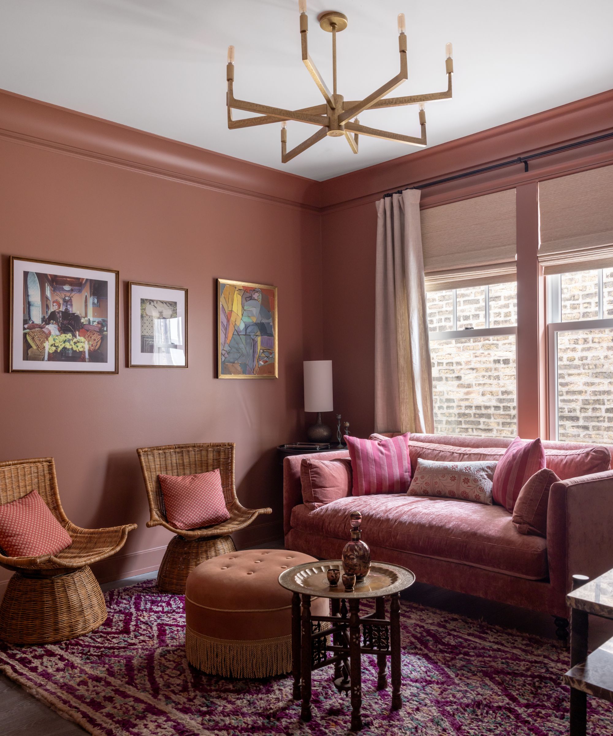 A small living room with terracotta walls, a dark pink rug, a pink sofa, two wicker chairs with pink cushions.