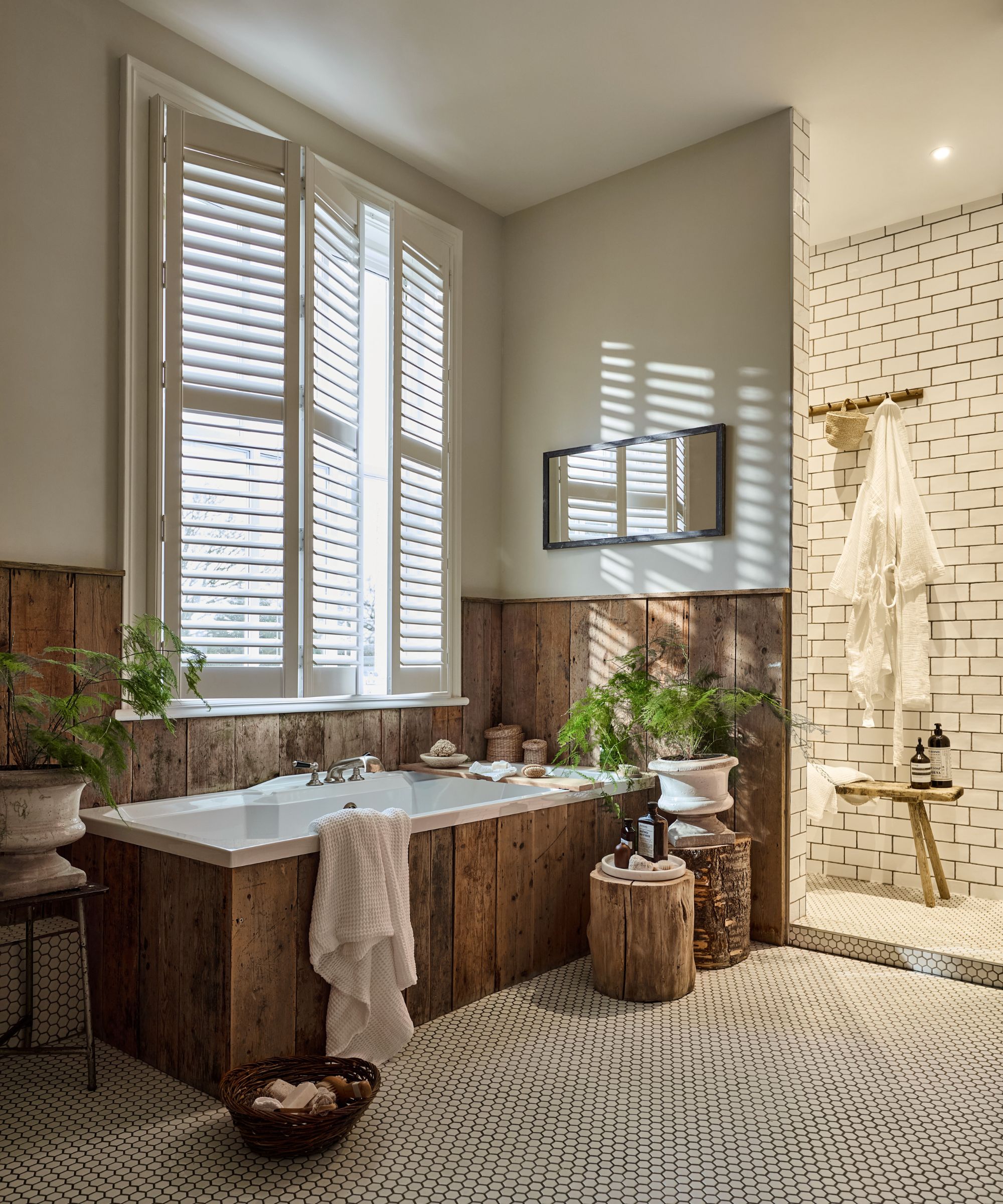 A warm, rustic bathroom with a wooden-panelled bath surround, white plantation shutters, and subway-tiled shower. Natural textures, ferns, and soft lighting create a serene, spa-like feel.