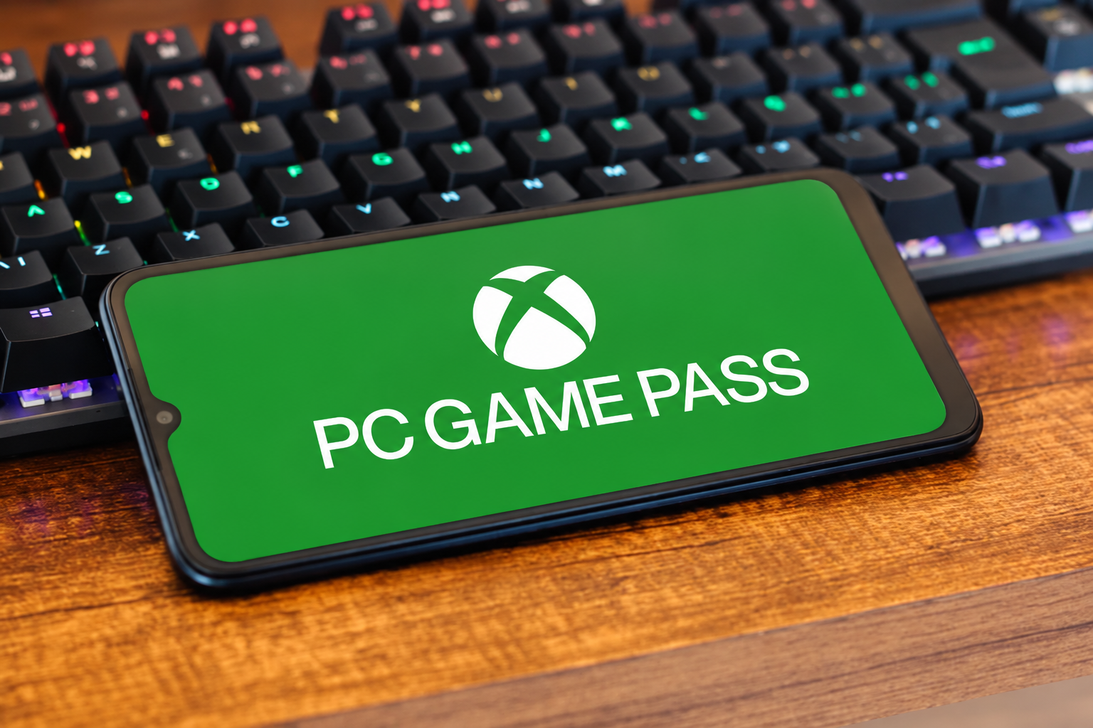 Smartphone displaying the Xbox PC Game Pass logo on a green screen, resting diagonally on a laptop keyboard illuminated by blue backlighting.