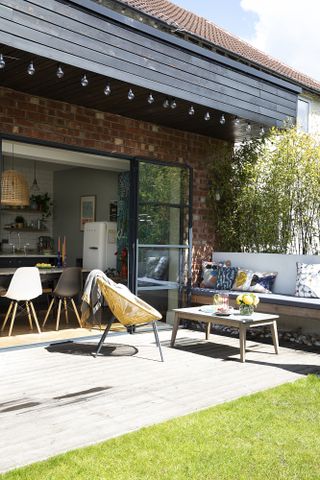 Decked area outside a rear extension with festoon lights, built-in bench, yellow string chair and wood coffee table