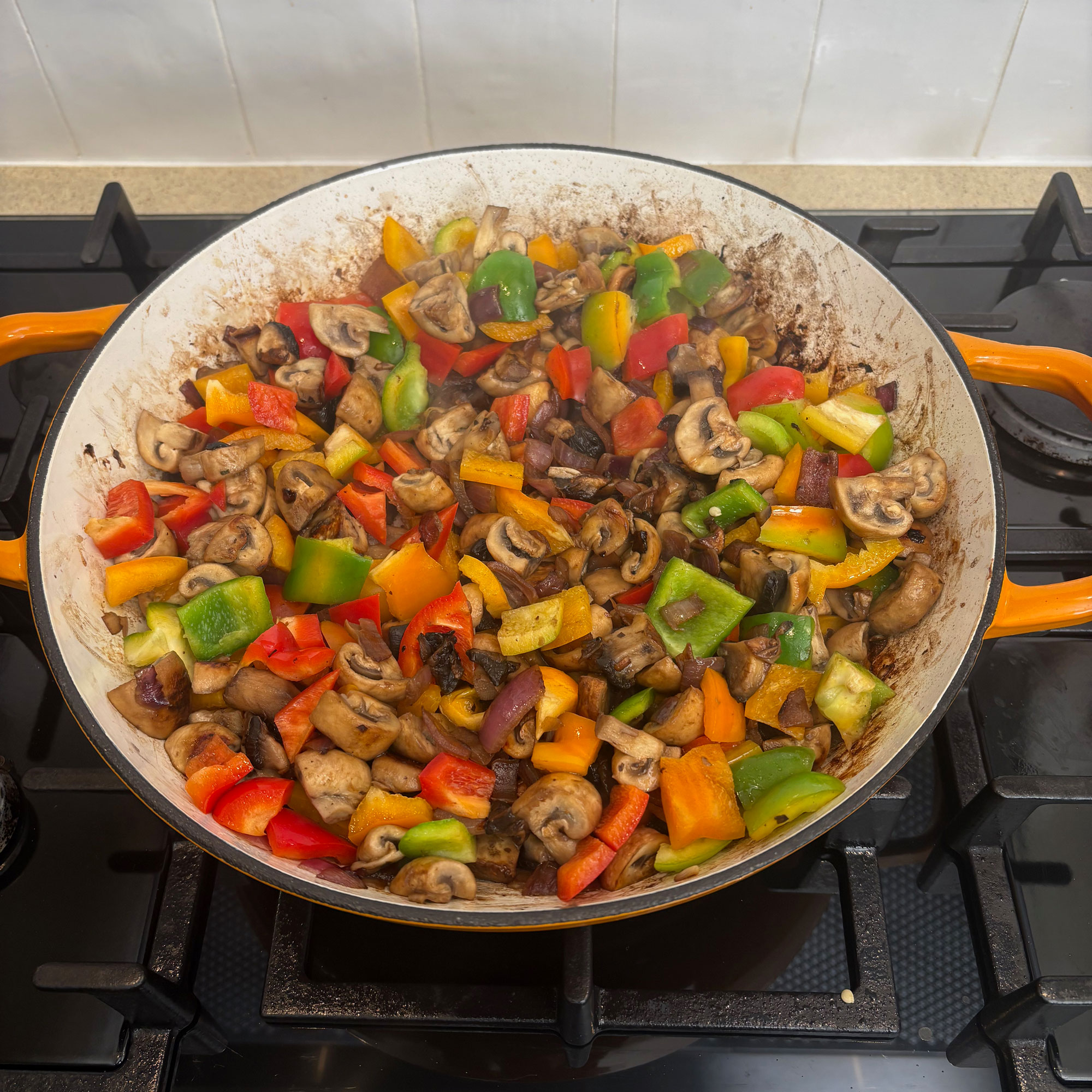 Orange cast iron pan on black hob with stir fried peppers, mushrooms inside.