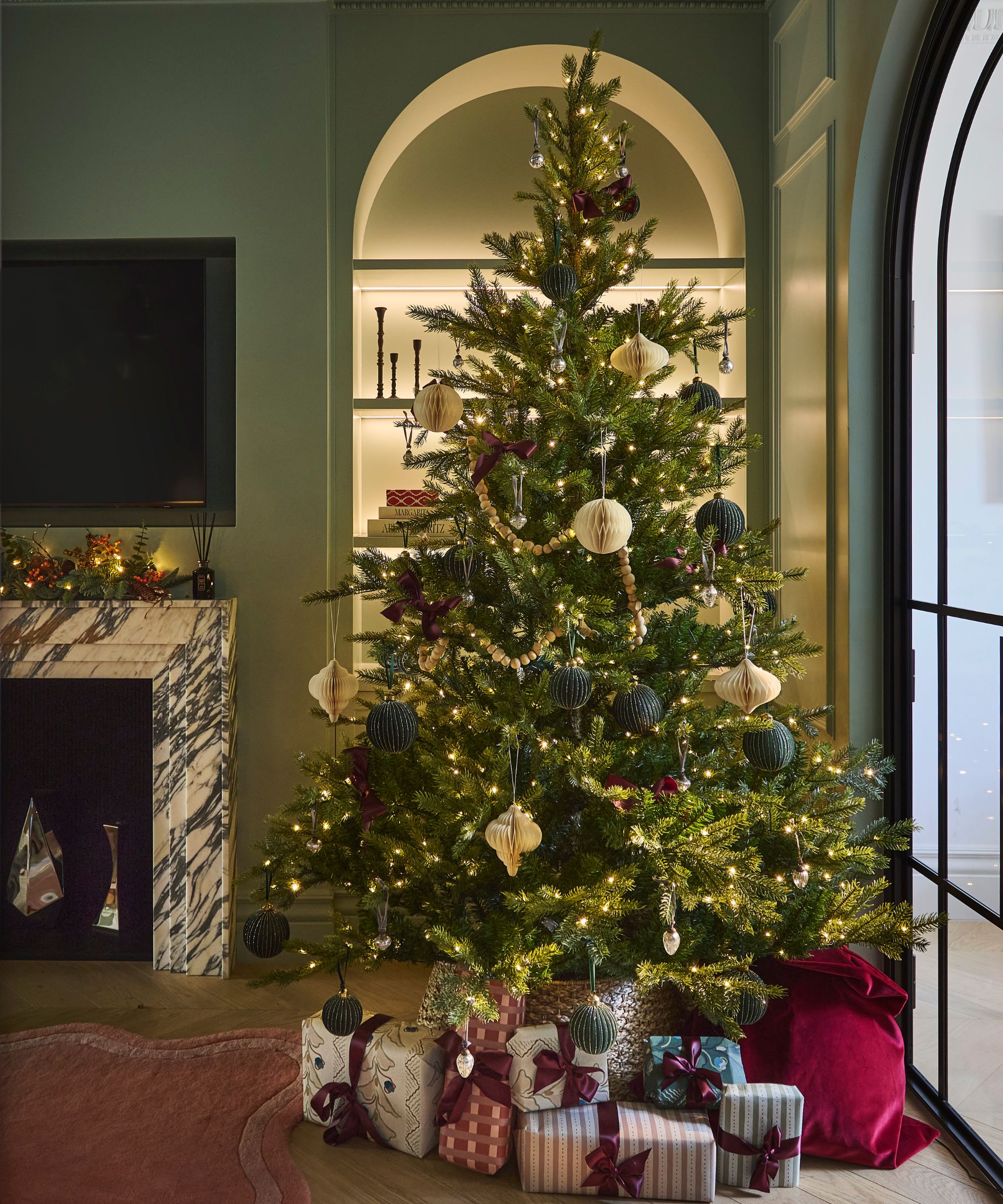 a muted green victorian living room with a marble fireplace, TV, arched bookcase all decorated for Christmas with a large lit tree and wrapped gifts