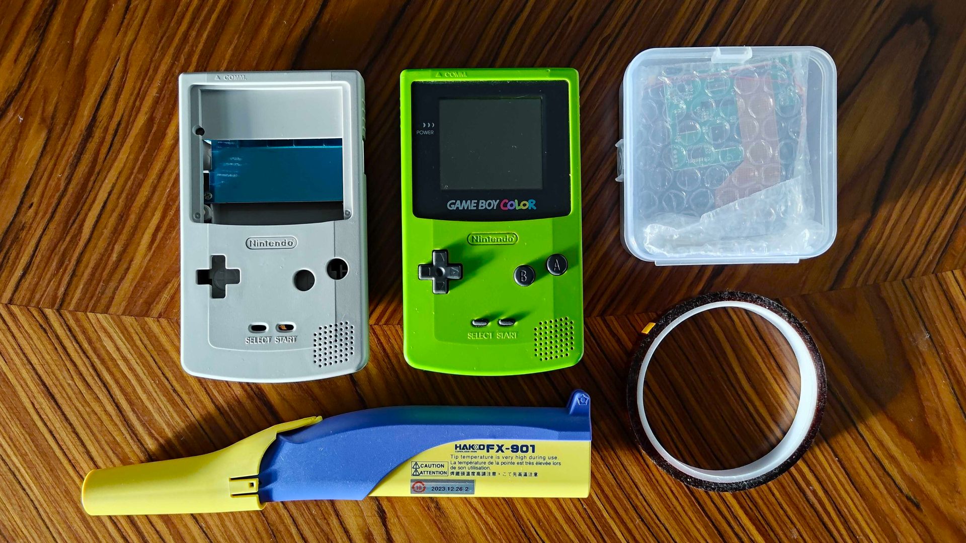 White Game Boy Color shell next to green console, Hakko soldering iron, kapton tape, and plastic box with AMOLED screen inside