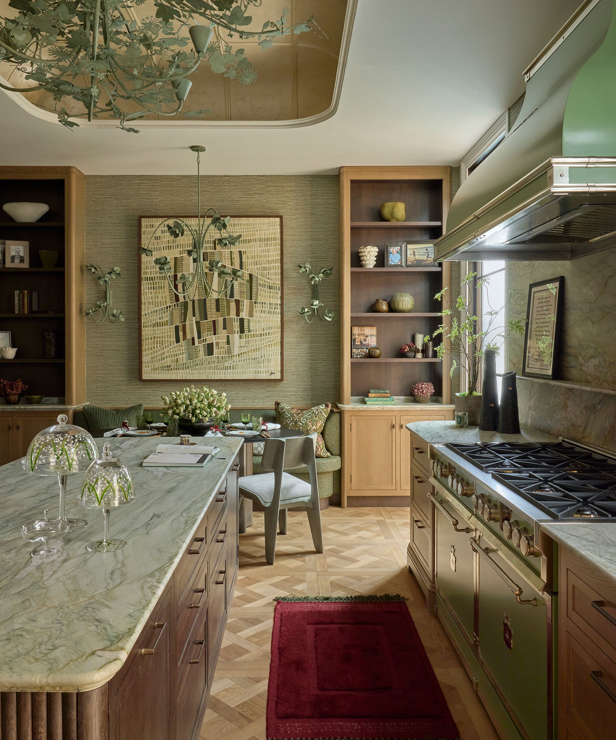 Kitchen with green marble island, parquet wooden floor, a green range oven, a red rug, and grasscloth walls with large wooden shelving units and wrought iron floral pendant lights