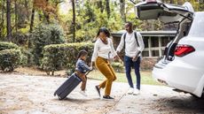 A toddler rides a suitcase that his mom is pulling toward the car while his dad looks on.